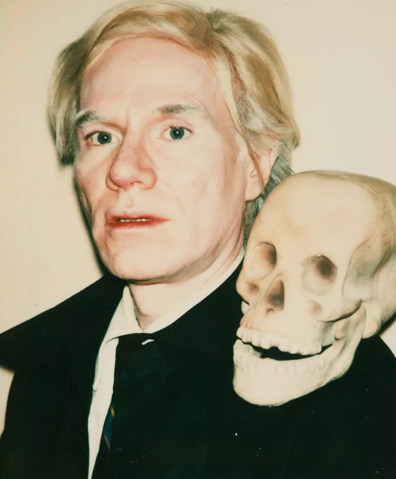 a photographic portrait of Andy Warhol, looking pensive, with a human skull perched on his left shoulder